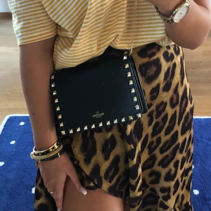 Äkta Valentino väska  - En Valentino väska, i svart med guld detaljer. Använd fåtal gånger så i väldigt bra skick. Nyköpt för två år sedan på NK för 9.899kr. Den ser liten ut men får plats med mobil, plånbok & lite till!  