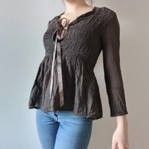 Mörkbrun blus, med sidenrosett framtill - Mörkbrun blus i bomull med sidenband att knyta framtill. Tyget är smockat (stretch) över bysten och nedanför faller det som volanger. På sista bilden kan ni se en urringning/glipa som blir under rosetten när man knyter sidenbanden. Passar XS