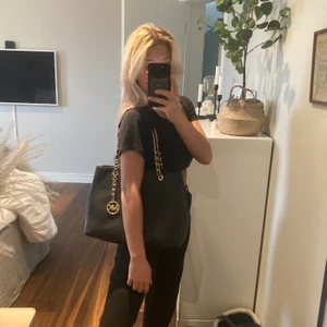 Michael Kors väska - Michael Kors väska i klassisk modell med gulddetaljer. Super fint skick utan några repor. Köpt för ca 4000/5000kr. Mycket rymlig väska som rymmer A4 papper, block och böcker. Två delar med ett fack i mitten som stängs med dragkedja. Fyra innerfickor och ett fack med dragkedja. Väskan stängs med en magnet. Väskan har även fyra guldknoppar på undersidan som fötter för att förhindra repor. 