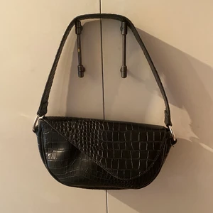 Svart axelväska  - Svart axelväska med krokodilmönster, köpt från Nelly. Väskan stängs med en knapp har ett bredare axelband. Finns även ett litet innerfack med dragkedja. Väskan är i fint skick och jag säljer den för att jag inte använder den längre. Frakt: 66kr spårbart 🖤  