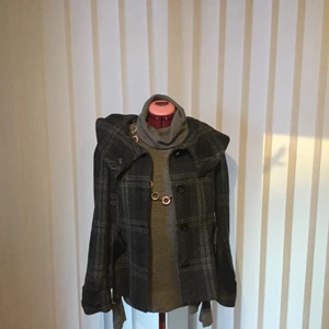 Kort jacka med kapuschong - Snygg kort jacka med kapuschong. Färg grå svart med glitter effekt. Kapuschongen är samlad med ett band så den ligger fint draperad på jackans rygg. Försedd med konstgjort skärp. Vanlig knäppning med trycknknapp och fyra svarta knappar. Material acryl, ull och polyester. Tvättråd 30 grader Strykes med svag värme. Svart foder. Längd 62 cm ärmlängd 61 cm mått från vänster axel till höger axel 41 cm. Tar swish