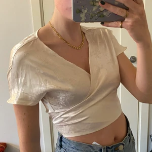 Omlott-topp i satin - Jättesöt omlott-topp i ett glansigt material & fint mönster. Vit-beige med lite ljusrosa ton. Säljer då den inte kommer till användning. Fint skick. Storlek S/36 men går att anpassa med tanke på knytningen