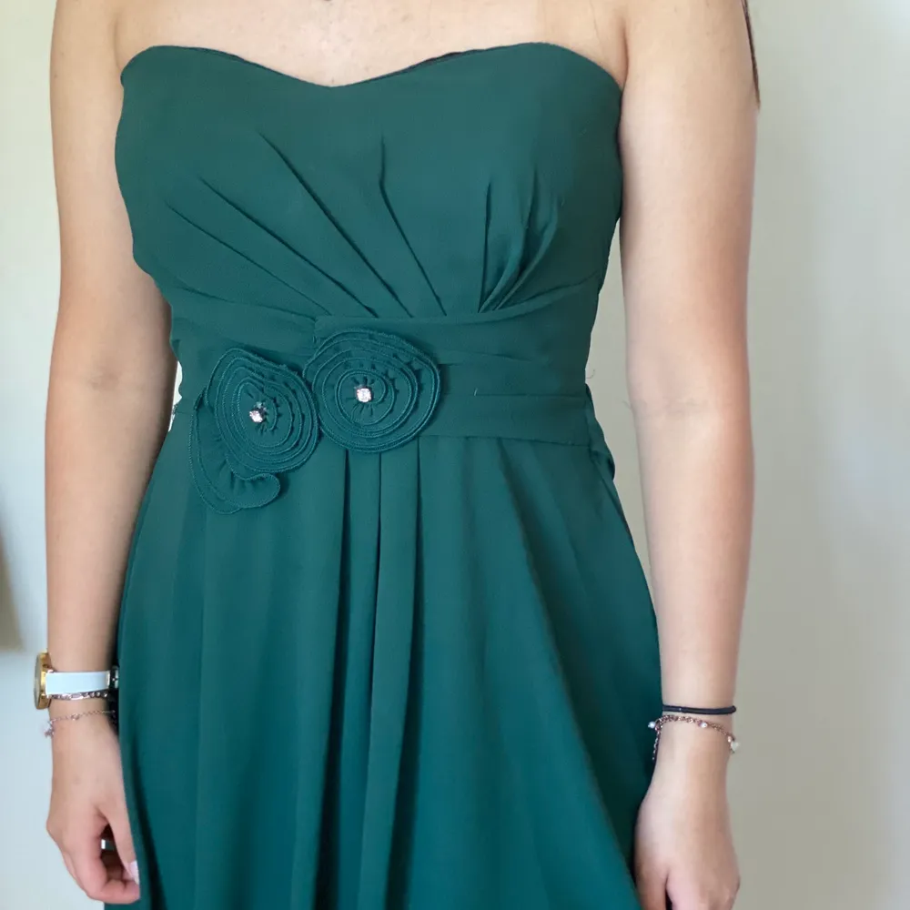 En kort of shoulder klänning i en mörkgrön färg. Runt midjan finns som ett bälte med ’blommor’ på. Använd mycket men i gott skick. Klänningar.