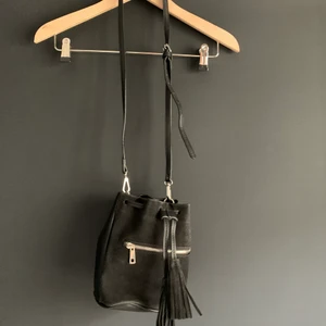 Bucketbag i läderimitation - Liten bucketbag i mocka och läderimitation. Yterfack med dragkedja och band med tofsar. Svart färg. Fint skick. Köpare står för frakt.