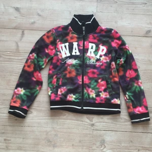 Färgglad flise. - Detta är en färgglad flise i ett tunt material. Den har en svart krage med en vit rand på. Samma längst ute i ärmarna är det samma material som på kragen och samma färg.