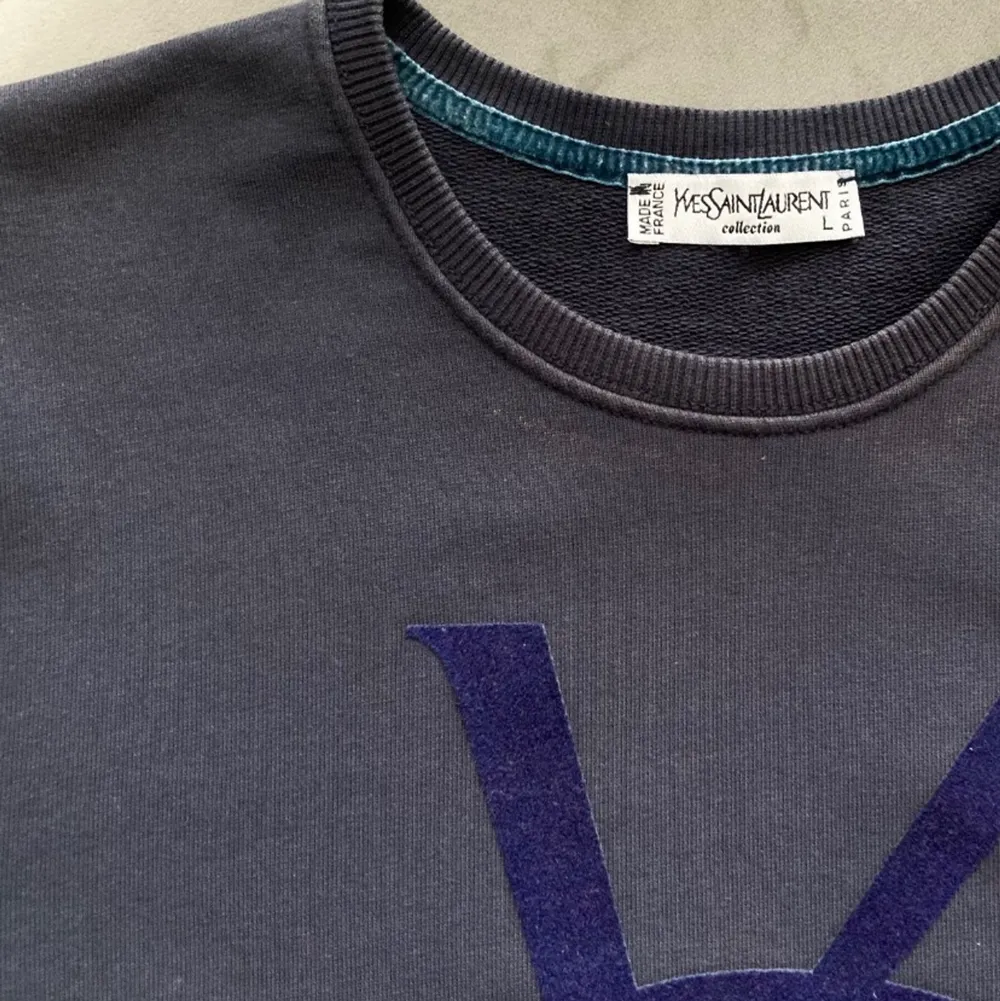 Mörkblå sweatshirt från YSL med stor logo i flock print. Storlek large men passar även medium. Tröjan är överlag i bra skick men det finns två defekter. Svaga fläckar efter blekning uppe vid kragen (bild 2) och viss slitning längst ner på logon (bild 3). . Hoodies.