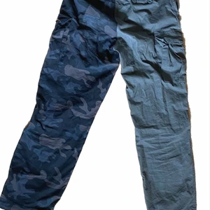 Tvådelade cargobyxor oliv/mörk camo - Tvärfeta cargobyxor med olika färg/mönster på varje ben. Ena benet är olivfärgat, andra har ett mörkt camoflage mönster. 40 cm i midjan, 80 cm långa. Sytt av mig av två par brandit bdu ripstop byxor. Aldrig använda.