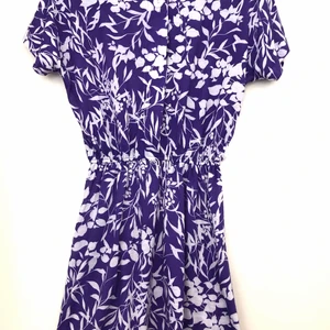 Lila klänning med vita blommor. Retro. - Retro klänning med lite markerad midja och axelvaddar. Knappar upptill. 100% polyester.