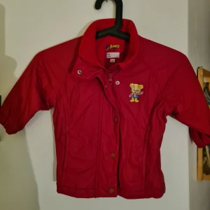 Bamse barn jacka storlek 80 - Röd Bamse jacka för en liten tjej. Storlek 80. Finns både dragkedja och knäppning.  På baksidan av ena ärmen finns en vit "prick' ca 2,5 cm.( ser ut som en liten tippex fläck) samt ett litet hål på samma ställe. Vid intresse kan jag sända bild på detta. Pga att jackan har ett litet hål samt en vit prick jag inte vet om det går att tvätta bort säljer jag ut den för endast 60 kr.  För övrigt i fint skick #bamse by lindex 