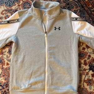 Under Amour långärmad tröja med dragkedja  - Långärmad Under Armour grå tröja med vita detaljer & dragkedja. Använd en gång. Skön insida i form av tunn fleece. Storlek anges ej, men    för barn ca 150 cm