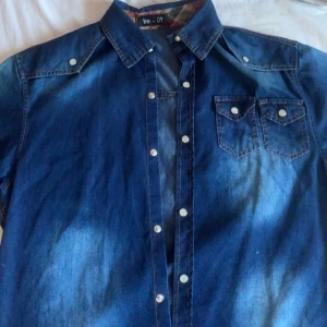 Kortärmad jeansskjorta  - supercool jeansskjorta med coola detaljer. har ingen aning om vart den är köpt då vi haft den jättelänge bara att ingen använt den så den är i nyskick! bara att skriva vid fler funderingar!