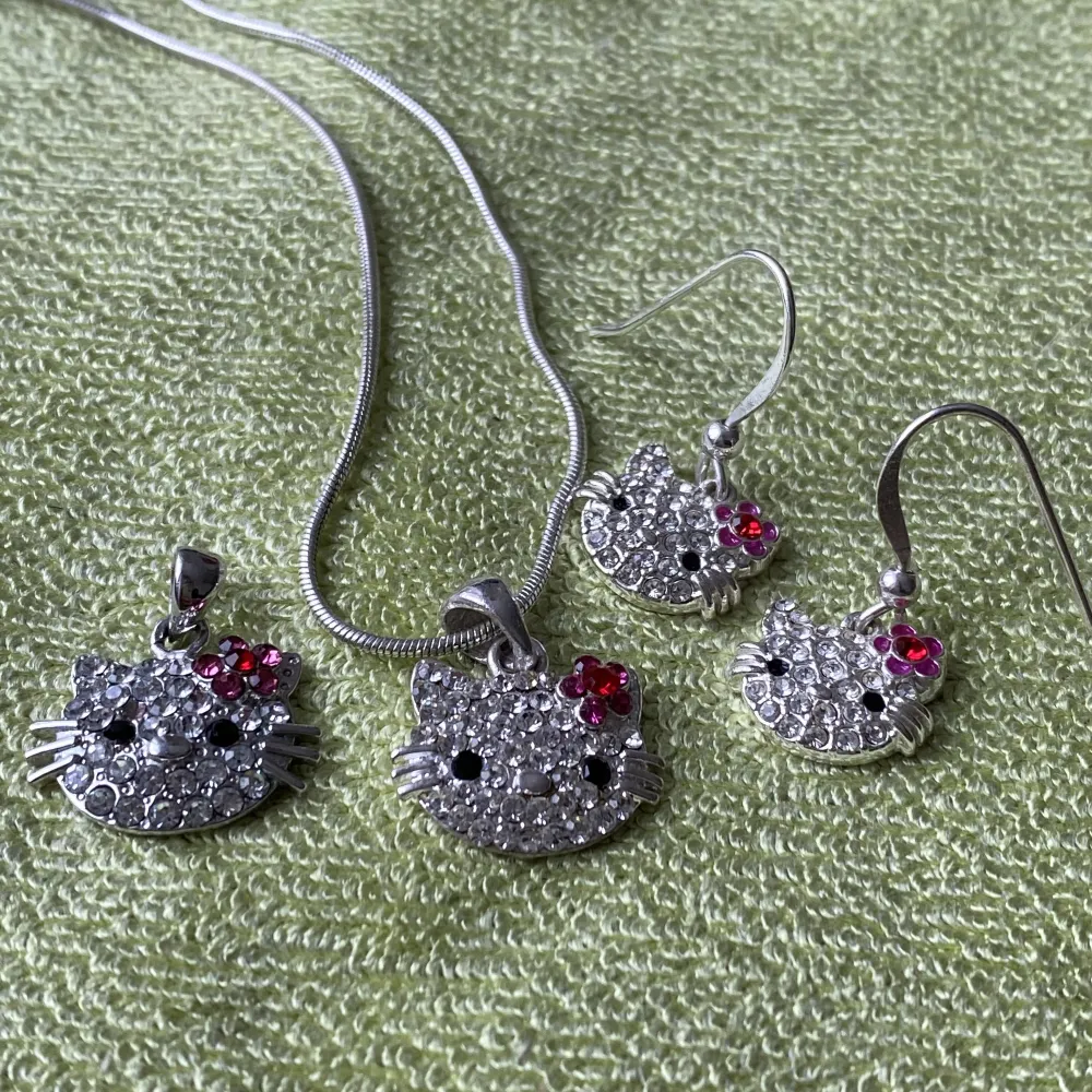 Set med smycken föreställande Hello Kitty med strass-stenar. Ett halsband, ett par örhängen och en extra berlock. Halsbandet är som kortast 20 cm och längst 26 cm runt halsen.  Berlockerna är 1.2-1.6 cm breda.  Möts upp eller fraktas spårbart.. Accessoarer.