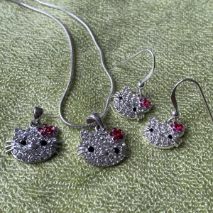 Hello kitty smycken - Set med smycken föreställande Hello Kitty med strass-stenar. Ett halsband, ett par örhängen och en extra berlock. Halsbandet är som kortast 20 cm och längst 26 cm runt halsen.  Berlockerna är 1.2-1.6 cm breda.  Möts upp eller fraktas spårbart.