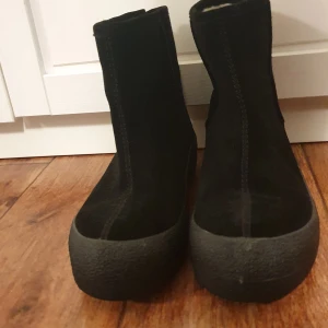 Svarta curlingboots i mocka - Svarta curlingboots i mjuk mocka och ullfoder. Guldfärgad dragkedja baktill. Uttagbar innersula i ull. Sulhöjd ca 3,5 cm. Strl 40. Endast gått med dem inomhus. Inköpta 2022 för 700 kr.  Endast gått med dem inne. Säljes pga att de tyvärr inte passar min