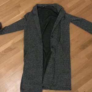 Tunn mörkgrå kappa  - Tunn mörkgrå kappa i fint skick, använts endast ett fåtal gånger. Passar till våren. Fickor. Elegant och stilren 