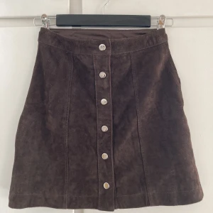 Mörkbrun kjol - Snygg minikjol i mörkbrunt mocka-läder. Knappar längst framsidan. 
