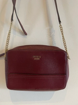 Handväska Guess - Snygg handväska i oxblodsfärgat/vinrött skinn från Guess. Justerbar axelrem i skinn och metallänk. Inköpt i Sverige och självklart äkta vara.  Mått: 24x16 cm  Väskan är i nyskick, endast använd 2ggr 