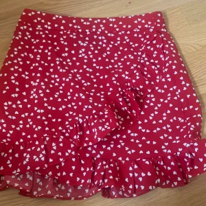 Röd kjol med volanger - Säljer denna söta kjolen då den är liten för mig. Har använt den 2 gånger och är i nyskick, kjolen är röd med vita hjärtan och harvvolanger kors över och längst ner.  Köparen står för frakten.