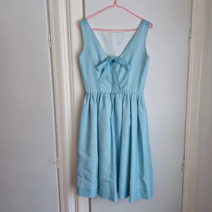 Vintage klänning ljusblå  - Fantastiskt fin klänning i ljusblå, stadigt tyg. Fodrad med silkigt tyg. Rosett och v-ringning i ryggen.  Nätt dragkedja.  Spännen under axelbanden för att hålla bh-banden på plats så att den inte syns (så himla smart!). En riktig 50-tals-pärla! 