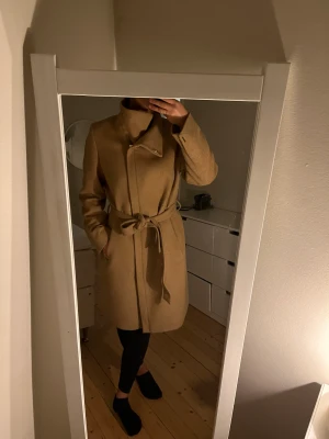 Beige kappa med ull - I nyskick, endast använd ett fåtal gånger. Kamel/ beigefärgad kappa från Esprit med dragkedja. Kappan är med ull och lite tjockare än en vanlig "tunn" kappa! Storlek L men passar M också