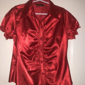 Kortärmad satin blus/skjorta - Säljer en kortärmad röd satin blus som är rynkad i midjan, vilket definierar midjan och får den att se bra ut. Blusen är i storlek 44 men är liten i storlek och motsvarar strl 40/42. I priset så är inte frakten inkluderad!!
