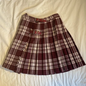 Rutig kjol - En vidröd pleated kjol i bra kvalitet 🤎 Köpt secondhand men i prima skick. Tjockt, varmt och tåligt tyg. Är som en wraparound (se bild 2) och fästs med bältena och en knapp på insidan. Går lite över knäna på mig som är 1,68