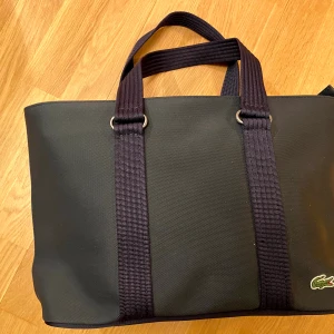 Ny Lacoste marinblå damväska  - Helt ny Lacoste marinblå damväska, vattentät med grönrandiga detaljer och fickor på insidan. Aldrig använd och i nytt skick. Från djur- och rökfri hem. Ord pris 1500kr Upphämtning Hägersten(Aspudden, Liljeholmen, Gröndal) alt Kungsholmen 