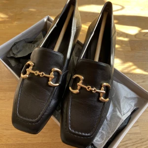 Loafers med gulddetalj - Säljer ett par Loafers i skinn från Tamaris i strl 37. Jag köpte dom på Boozt i april i år. Orginalpriset är 999kr men säljer dom för 500kr då de endast är använda max 3 gånger då jag inte tycker att de passar på mig. De är slutsålda och säljs inte l