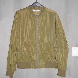 Grön bomberjacka i glansigt material - Säljer denna gröna bomberjacka i glansigt material. Den skiftar något i färg beroende på ljus men är normalt sett mörkt olivgrön. Den är i gott skick!