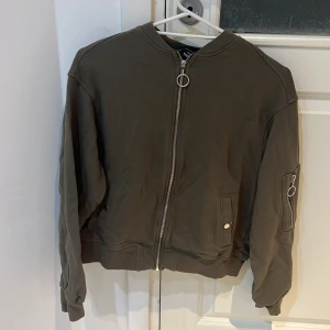 Tröja med dragkedja  - En khaki färgad tröja med dragkedja från KappAhl i storlek S. Den har även två fickor som stänger med knappar.