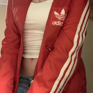 Röd Adidas  - En röd adidas jacka i unik modell i ett material som funkar för både träning och vardag. Luvan back går även att fälla ihop elle ha ute. Den sitter väldigt litet och croppat på mig som är 160cm och jag S/XS  Använd flertal gånger. Kolla bion för mer info