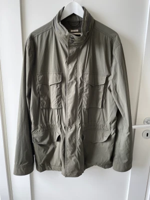 Safari jacka / M65 jacka - Väldigt fin safari/M65 jacka. Olivgrön. Klassisk model med modern passform. Stretch material gör den väldigt skön att bära. Flera fickor. Utfällbar huva i kragen. 