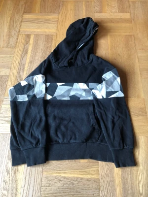 Långärmad tröja  - En svart långärmad tröja med luva på magen och ett svart/vit/grått marmor på bröstet och armarna. 