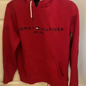 Tommy Hilfiger hoodie - En röd Tommy hilfiger hoodie i storlek herr M. Som går att ses på andra bilden så har tröjan en liten fläck.