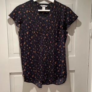 Marinblå H&M-blus - Marinblå blus med orange blommönster. Kort lite vid ärm över axeln. Sparsamt använd vid kontorsarbete. Snygg till kjol och byxor. 