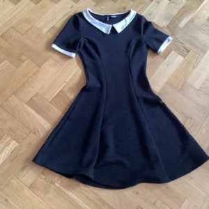Snygg klänning - Obs! Bortse från färguppfattningen på bilden, den är Svart! Snygg lite klockad klänning med vit krage och vita kantband på ärmarna. Stretchigt tyg, lite ribbad.
