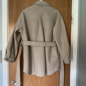 Zara jacka strl M - Säljer en beige skjortjacka med skärp från Zara. Den är i bra skick utom att en av sömmarna har gått upp lite på en av fickorna framtill (se sista bilden).   Nypris 559 kr, säljer för 150 kr. Hämtas i Sundbyberg eller skickas mot fraktkostnad.