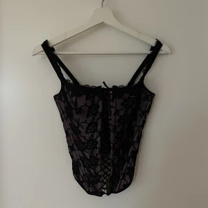 Svart och rosa/lila korsett i spets - Korsett med svarta och lila/rosa inslag. Spets, snörning framtill och hakar baktill. Återigen omöjlig att ta en rättvis bild på 
