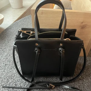 MINICITYVÄSKA MED KNUTAR FRÅN ZARA - Axelremsväska från Zara. Den är svart med knutar på sidorna och guldfärgade metalldetaljer, samt dragkedja. Axelremmen kan justeras och tas bort. Höjd: 16,2 cm Bredd: 18,5 cm Djup: 8 cm. Aldrig använd så är i nyskick. 