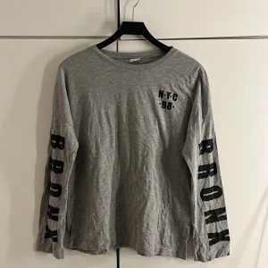 Långärmad grå tröja  - En långärmad grå tröja med tryck på ärmarna som säger ”BRONX” och ett tryck på vänstra delen av bröstet som säger ”N•Y•C” och under den står det ”•98•”. Den är använd få tals gånger och är i väldigt bra skick. 