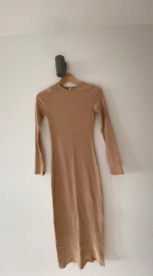 Beige tight klänning - Långärmad ribbad klänning. Beige i storlek XS. Ganska lång, går en liten bit under knäna på mig som är ungefär 165 cm lång. Slits på vänster sida. Den är använd 2-3 gånger max.  Köpt på Nelly. 