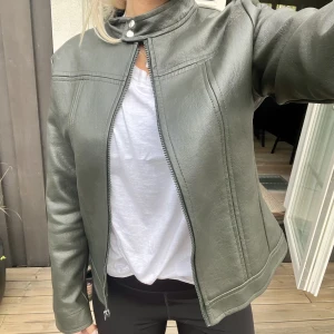 Mörkgrön skinnjacka  - Mörkgrön nästan svart/grå jacka i skinnimitation. Spänne vid halsen och dragkedja. Jackan har ingen storlek men skulle säga att den passar S/M beroende på hur man vill att den sitter! Bra skick men en liten reva på ryggen. 