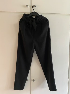 Kostymbyxor, paperbag waist strl 34 - Kostymbyxor med paperbag waist och omutbart skärp. Dragkedja och knapp med hake. Något croppade Inga synliga tecken på användning 