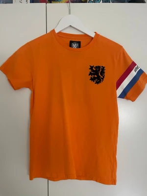 Orange fotbollströja med tryck - Snygg orange fotbollströja med svart lejontryck på bröstet och siffran 10 på ryggen. Ärmen har ränder i rött, vitt och blått samt texten 'Aanvoerder'. Perfekt för dig som gillar sportig stil och vill sticka ut på planen eller till vardags.