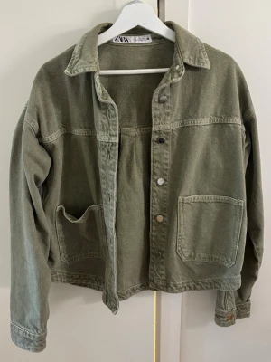 Populär jeansjacka från ZARA - Snygg olivgrön jeansjacka från ZARA med klassisk krage, tryckknappar och två stora fickor framtill. Jackan har en avslappnad passform och är tillverkad i slitstarkt jeanstyg. Perfekt för dig som gillar en cool och enkel stil.