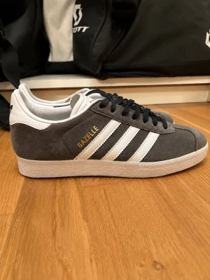 Adidas Gazelle gråvita sneakers - Säljer ett par klassiska Adidas Gazelle sneakers i grå mocka med vita detaljer och svarta skosnören. Skorna har de ikoniska tre ränderna på sidan och texten 'GAZELLE' i guld. Yttersulan är vit och ovandelen är i mjuk mocka för en clean look.