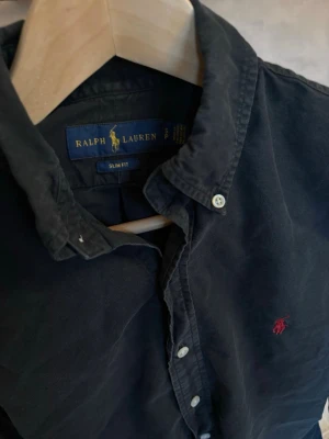 Svart skjorta från Ralph Lauren - Klassisk svart skjorta från Ralph Lauren med slim fit-passform. Skjortan har långa ärmar, knappar framtill och den ikoniska röda logobrodyr på bröstet.   Storlek 170/92A herr