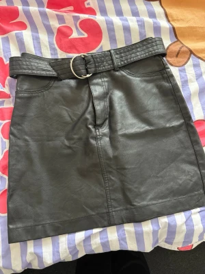Svart kortkjol i skinnimitation med bälte - Snygg svart kortkjol i skinnimitation med ett matchande bälte och silverfärgat spänne. Kjolen har fickor framtill och en rak passform som ger en cool vibe. Perfekt att styla med en stickad tröja eller t-shirt för en trendig look.Använd en gång 
