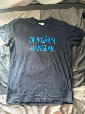 Blå Djurgår'n Manglar t-shirt - Mörkblå t-shirt med texten 'DJURGÅR'N MANGLAR' i ljusblått på framsidan och en stor etta i gult, rött och blått på ryggen. Under siffran står det 'DJURGÅR'N'. Perfekt för dig som vill visa ditt stöd för Djurgården. Klassisk passform och mjukt bomullsmaterial.