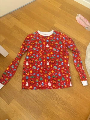 Röd julpyjamas med mönster - Säljer en långärmad pyjamastopp i rött med färgglatt julmönster som inkluderar tomtar, granar, paket och snögubbar. Tröjan har vita muddar vid ärmslut och halsringning. Perfekt för mysiga vinterkvällar och julfirande. Med ett par matchande byxor 